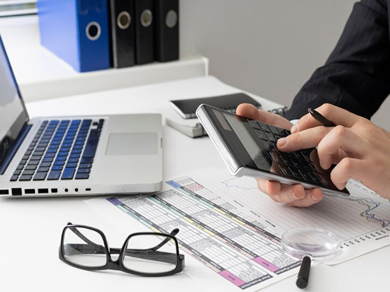 Person using a calculator at a desk with a laptop, analyzing financial charts and documents.